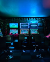 Dimly lit gaming arcade featuring a variety of brightly illuminated slot machines and video gaming screens. Neon signs and colorful displays create a vibrant atmosphere. The arrangement of gaming machines suggests an inviting space for entertainment, with chairs positioned for players.