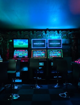 Dimly lit gaming arcade featuring a variety of brightly illuminated slot machines and video gaming screens. Neon signs and colorful displays create a vibrant atmosphere. The arrangement of gaming machines suggests an inviting space for entertainment, with chairs positioned for players.