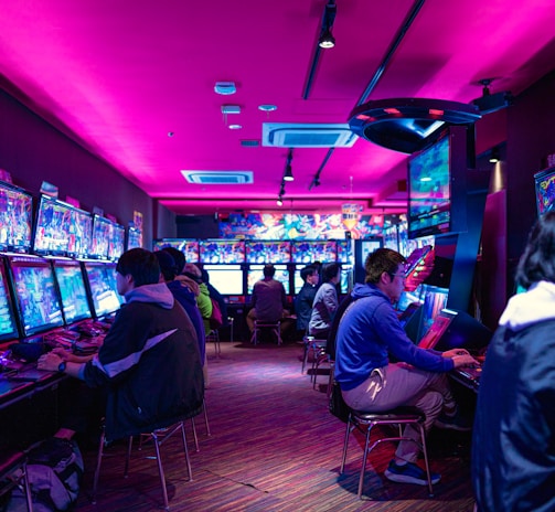 A vibrant arcade with rows of gaming machines adorned with colorful screens. Several people are immersed in playing games, seated on stools in a dimly lit room accented by an intense pink ceiling light. The atmosphere is lively and engaging.