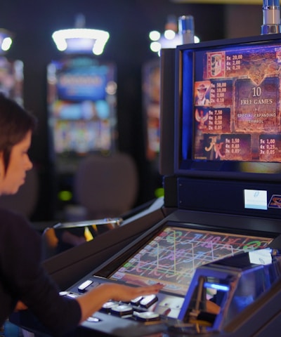 A person is seated at a slot machine console in a casino, focusing intently on the game displayed on the screen. The background shows several other illuminated gaming machines, adding a vibrant and colorful atmosphere to the scene.